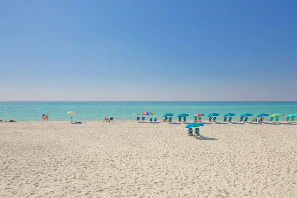 A vibrant beach scene featuring colorful umbrellas, families and children playing in the sand, and the clear blue ocean stretching out along a pristine shoreline. The atmosphere is lively and inviting, with groups building sandcastles and engaging in beach games.