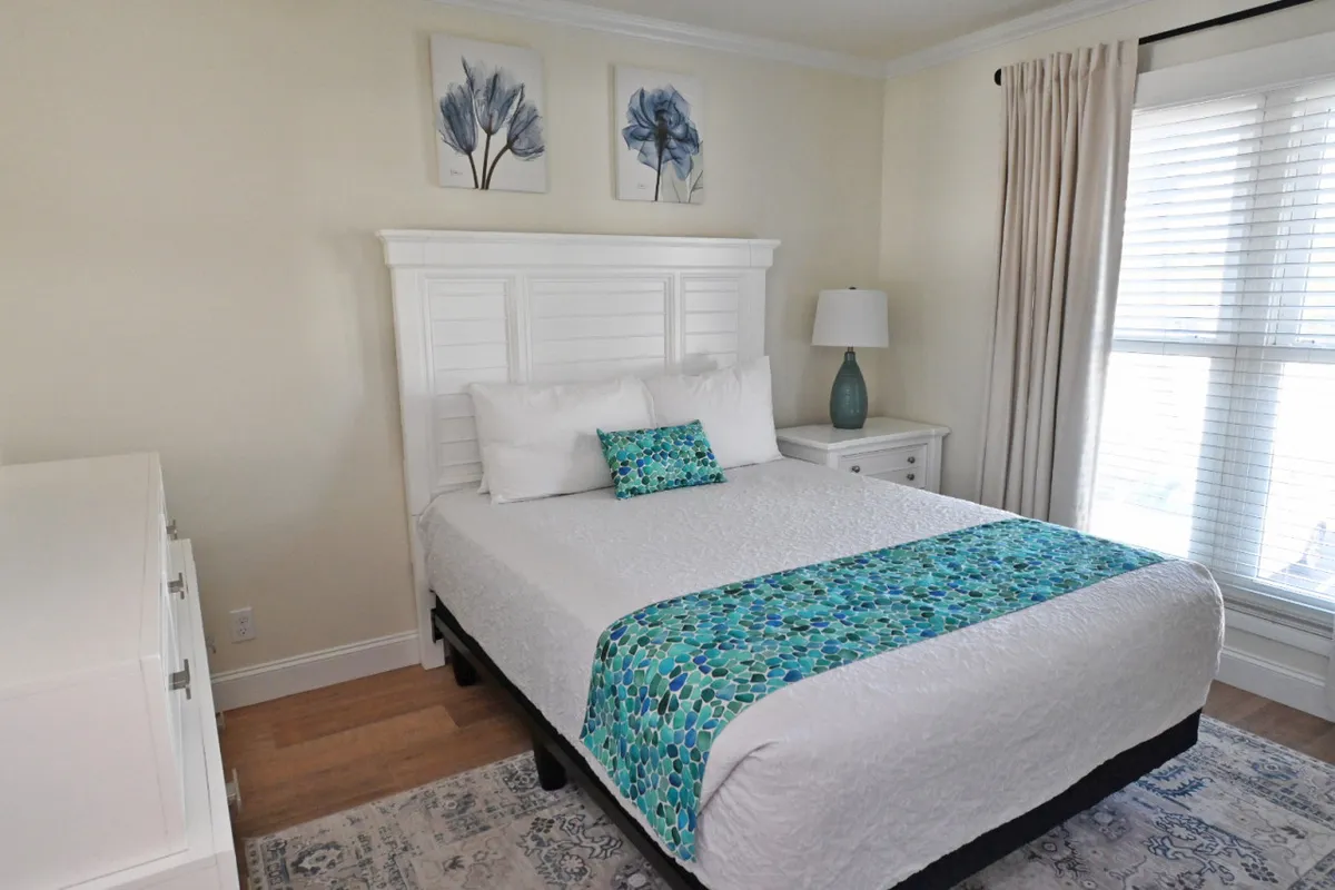 A serene bedroom scene showcasing a bed adorned with a decorative throw, a nightstand with a softly-lit lamp, and tasteful artwork on the wall. Natural light filters into the room, highlighting the tranquil color palette and inviting textures.