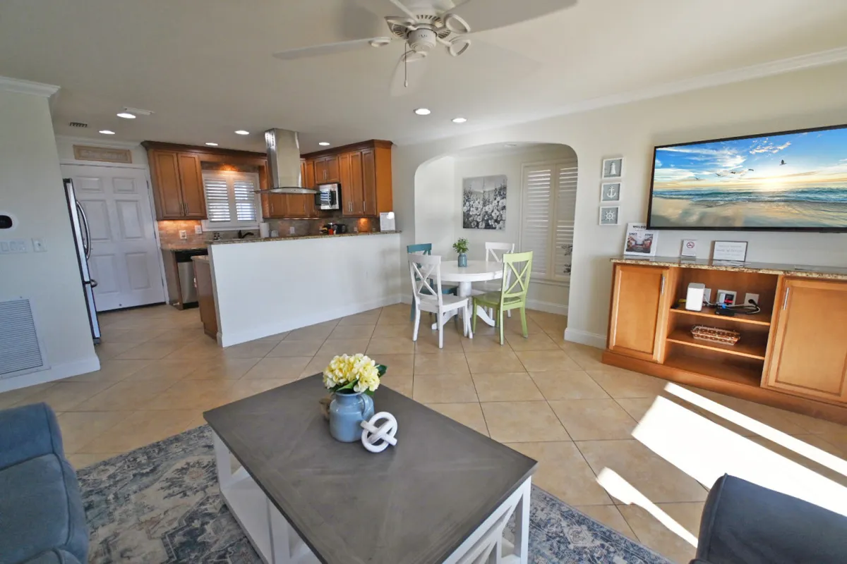 This image presents an open-concept living and dining area featuring a cozy sofa, a dining table with green chairs, and a television displaying a serene beach scene. Decorative touches like a coffee table with flowers add a touch of hominess. Natural light fills the space, creating a welcoming, relaxed vibe ideal for family gatherings, movie nights, or quiet escapes.