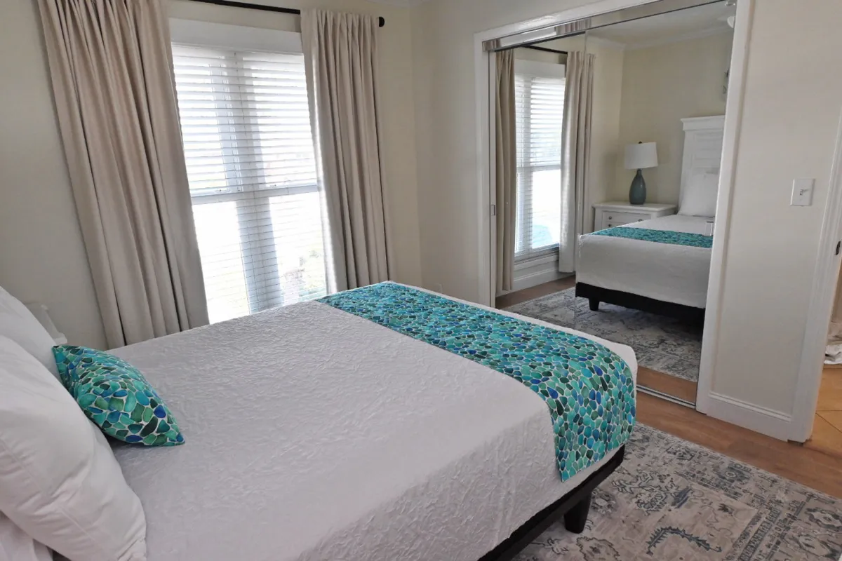 A bright, serene bedroom featuring a bed with a colorful throw, large windows allowing ample natural light, and a mirrored closet offering generous storage. The overall atmosphere is calm and inviting, ideal for unwinding after a busy day.