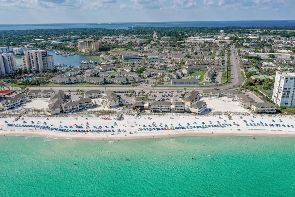 A lively beachfront scene featuring blue umbrellas, crystal clear water, and residential buildings in the background. The image exudes a relaxed, sun-soaked mood, with visible opportunities for water sports, swimming, and sunbathing.
