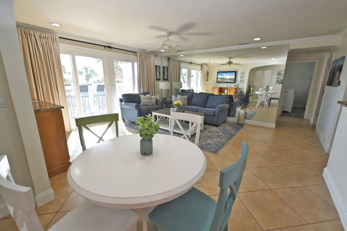 A bright, inviting interior featuring a dining table with colorful chairs, a plush blue sofa with decorative cushions, and large windows that fill the space with natural light. The layout is perfect for family meals, friendly gatherings, or simply enjoying a good book in a cozy atmosphere.