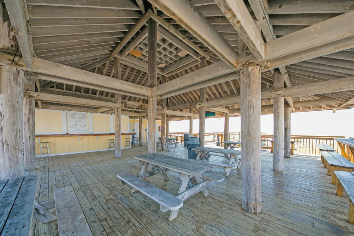 An open-air pavilion features sturdy wooden picnic tables and beams, with breezy views connecting to the outdoors. The communal space is ideal for gatherings or sunset barbecues, inviting a sense of togetherness and relaxation.