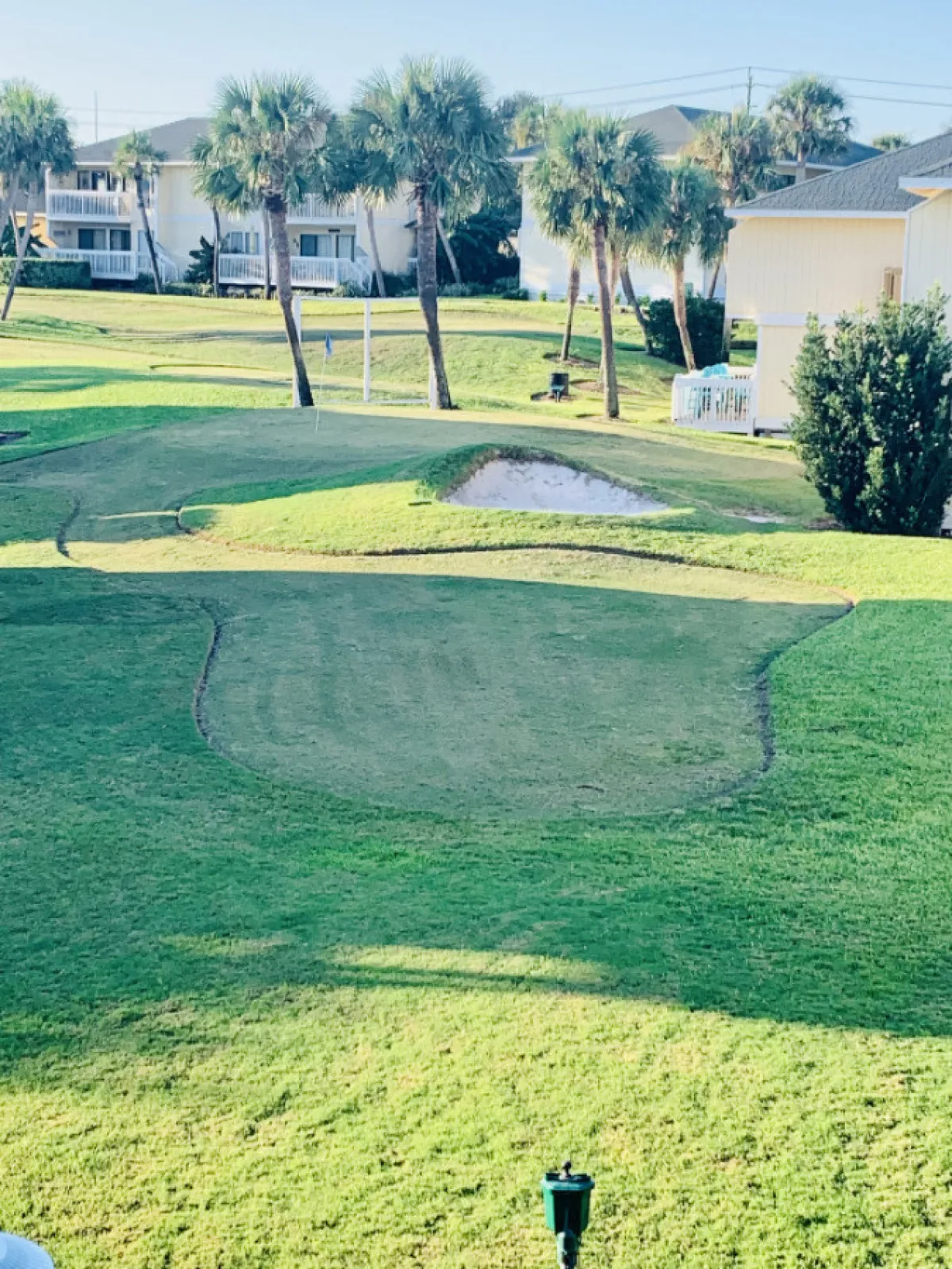 This image features a lush green golf course lined with palm trees, a sand bunker, and residential buildings in the background. The setting evokes a relaxed, resort-like atmosphere and highlights an area designed for both active play and scenic enjoyment.