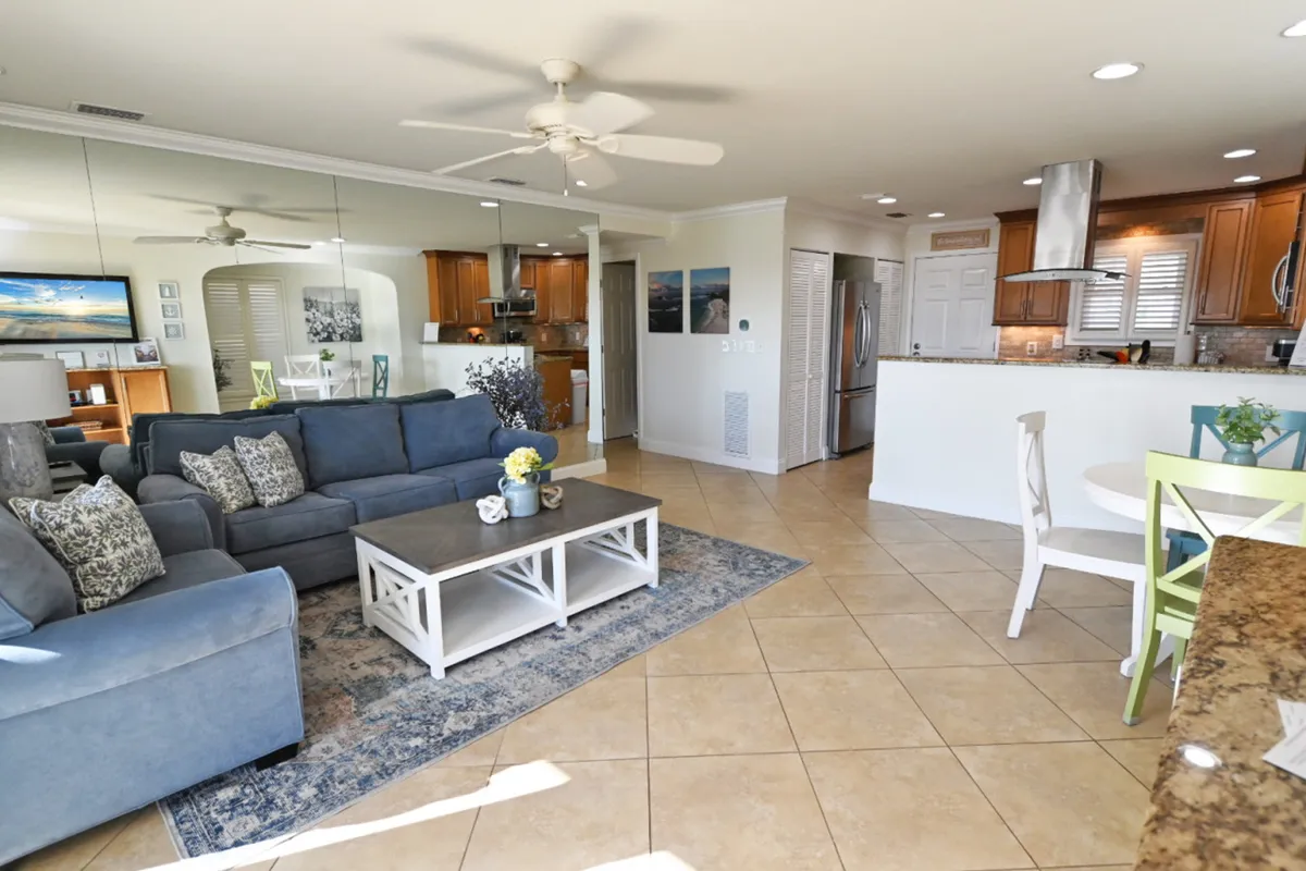 The image showcases a bright, open-concept living space with plush blue sofas, a crisp white coffee table, a stylish dining area with colorful chairs, and a sleek modern kitchen featuring stainless steel appliances. Sunlight fills the room, creating a warm and inviting atmosphere perfect for relaxing or entertaining after a day at the beach.