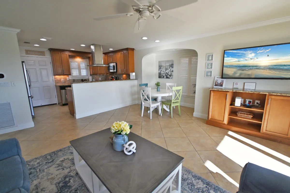 This image captures an open-concept living, dining, and kitchen area. The kitchen features stainless steel appliances and a hood, blending into a dining area with colorful chairs and a bright living space. The coffee table with a decorative vase and the large TV displaying a beach scene add warmth and a welcoming touch.
