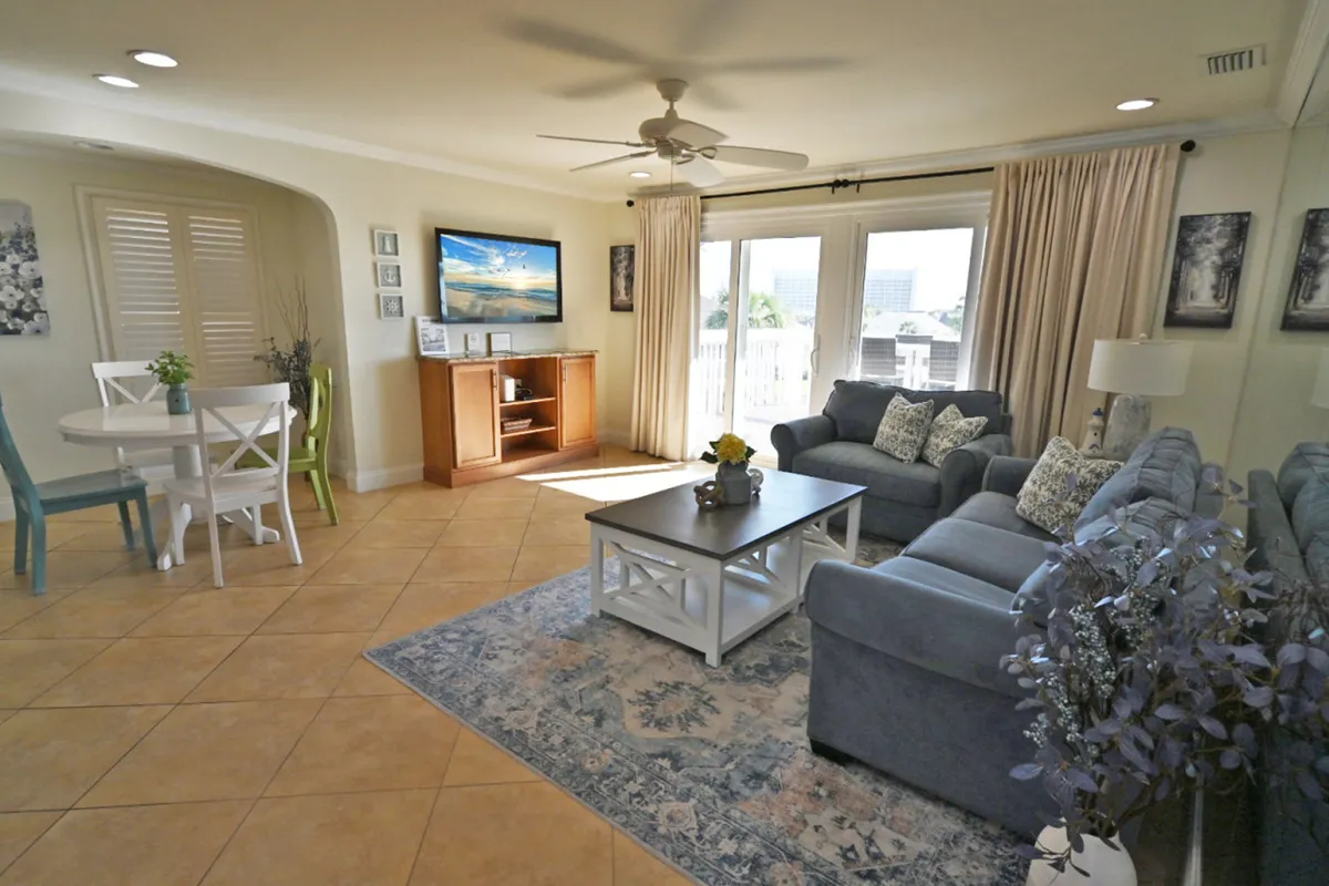 A comfortable living room with two gray sofas adorned with patterned pillows, a coffee table with a centerpiece, and a flat-screen TV mounted above a wooden entertainment unit. Glass doors lead to an outdoor balcony, and natural light fills the space, creating a welcoming, relaxing environment.