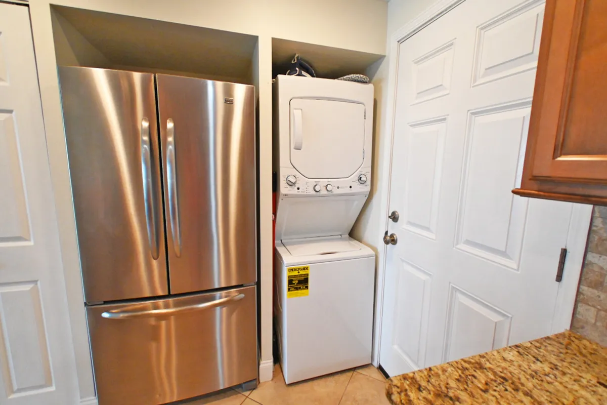 A practical corner of the home showing a stainless steel refrigerator, stacked washer and dryer, granite countertop, and a white door. The space is efficient and reflects thoughtful amenities for everyday vacation needs.