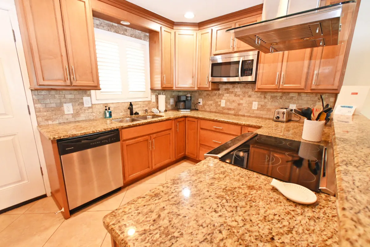 A close-up view of the kitchen, highlighting granite countertops, wood cabinetry, stainless steel appliances, and a window with white blinds. The scene feels homey and practical, emphasizing the kitchen's role as the heart of the home.