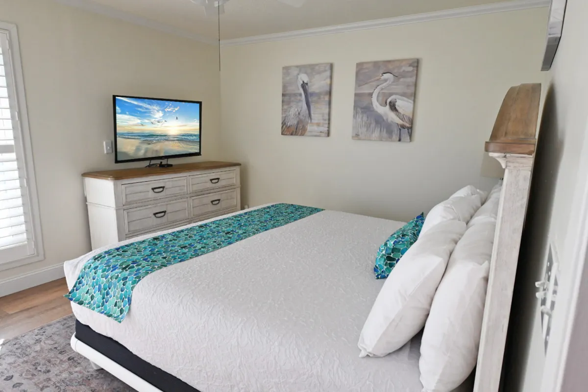 A tranquil bedroom with a large bed dressed in white and turquoise bedding, a dresser topped with a flat-screen TV displaying a serene ocean scene, and wall art featuring coastal birds. The overall ambiance is soothing and restful, with a focus on comfort and escape.