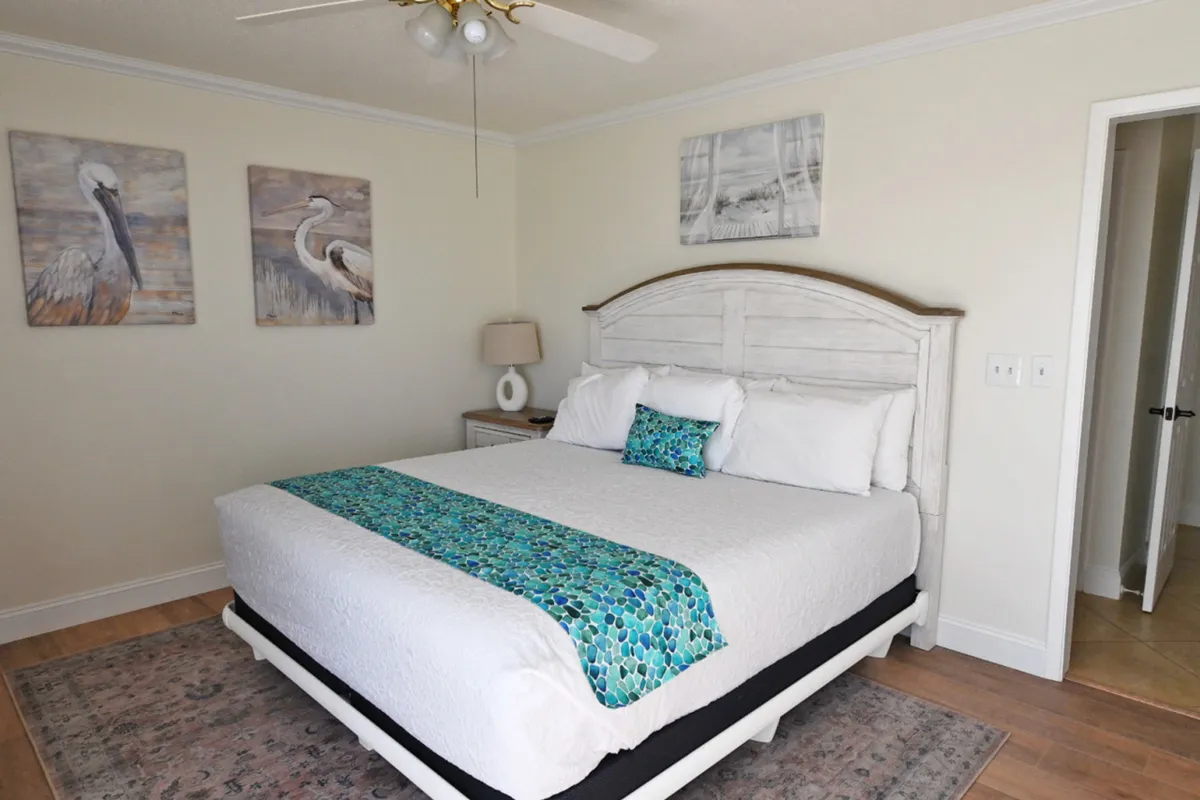 A spacious bedroom featuring a large white wooden bed with crisp white linens and a blue-green decorative runner and pillow. The room is accented with coastal-themed wall art depicting pelicans and beach scenes, a modern nightstand with a lamp, and a ceiling fan overhead. The calming color palette and soft lighting create a serene, inviting retreat.