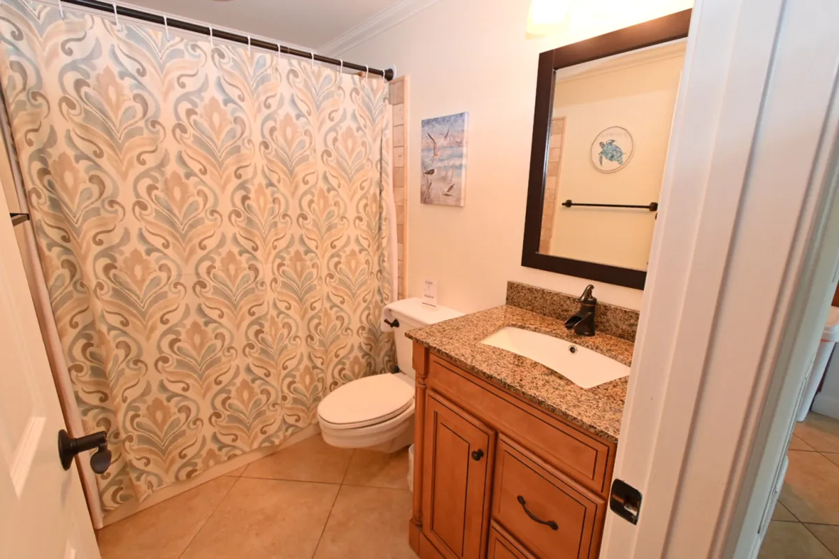 A contemporary bathroom featuring a patterned shower curtain in soft blue and beige tones, a granite countertop with a built-in sink, a wooden vanity cabinet, and beach-themed wall art. The space is bright, clean, and accented with subtle coastal charm.