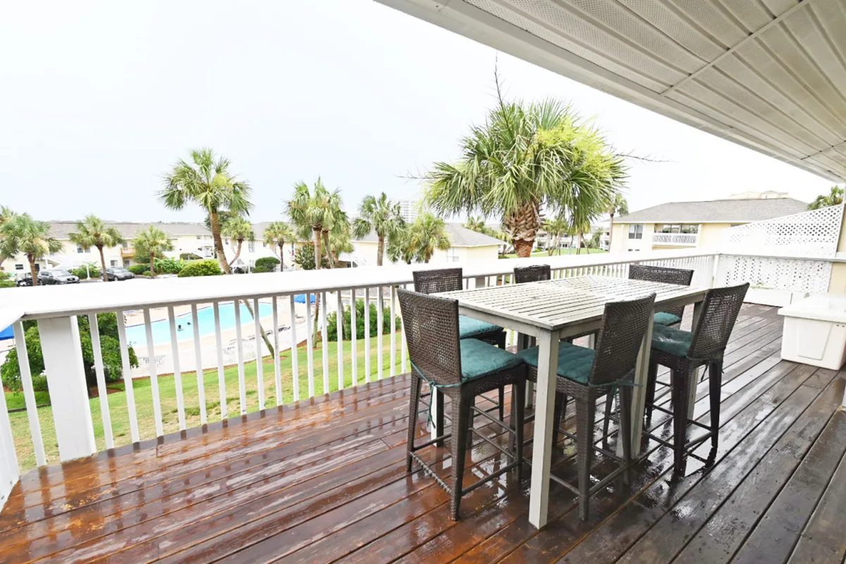 A wet wooden deck with a high dining table and chairs overlooks a sparkling heated community pool, surrounded by tall palm trees and tropical landscaping. The white railing frames the view, and the fresh look after a rain adds a sense of calm and renewal.