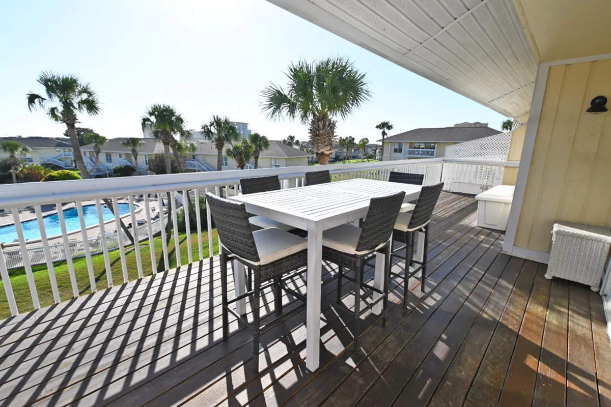 A spacious wooden deck with a white outdoor dining table and six woven chairs provides a comfortable spot to enjoy meals or drinks while overlooking the heated community pool below. Palm trees are scattered around the resort, creating a tranquil, vacation-like atmosphere.