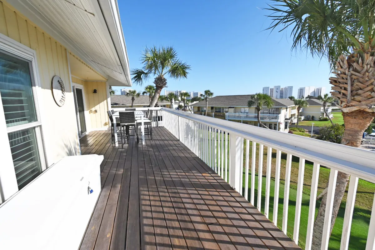 An extended wooden deck with white railings and an outdoor dining table is surrounded by lush palm trees, offering distant views of residential buildings and clear blue skies. The scene is flooded with natural light, highlighting the inviting, open-air environment.