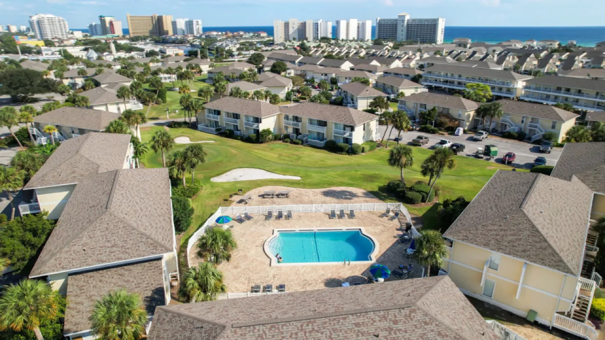 A central heated pool surrounded by lounge chairs, with multi-unit resort buildings, a meticulously maintained green golf course, and the ocean visible in the distance. The atmosphere is vibrant, suggesting ongoing activities like swimming, golfing, and socializing.