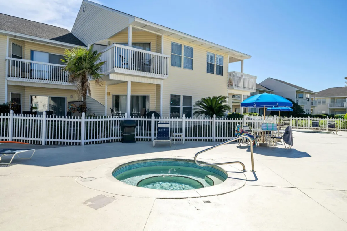 A bubbling circular hot tub is enclosed by a white picket fence within a landscaped pool area. Blue umbrellas shade patio tables and chairs, and two-story resort buildings with balconies overlook the inviting space.