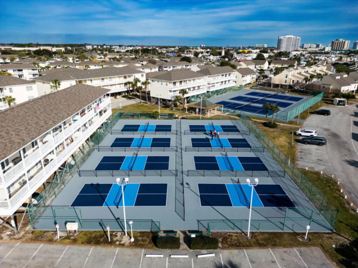 Multiple tennis and pickleball courts with players engaged in a match, surrounded by green spaces and residential resort buildings. The courts are active, underscoring the resort’s energetic and communal atmosphere.