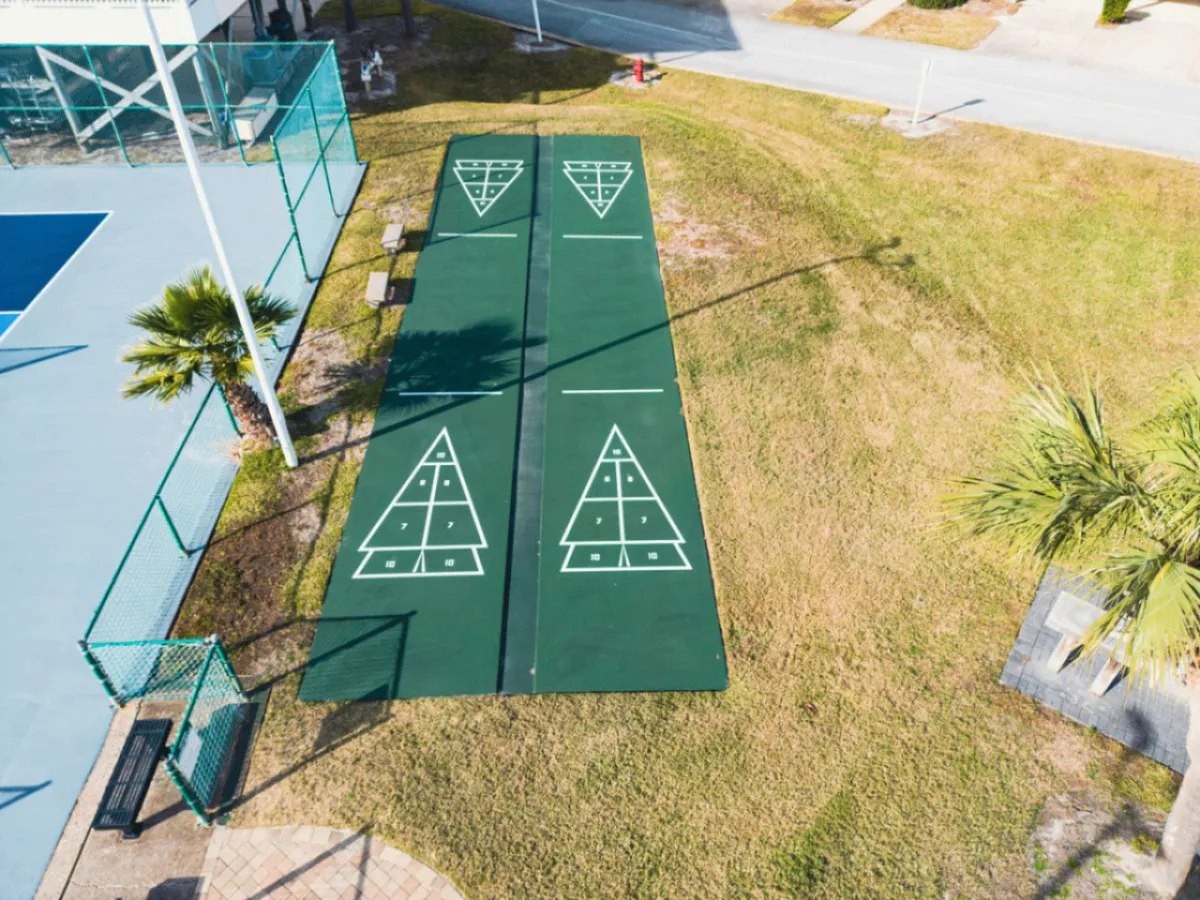 A bright outdoor recreation area featuring well-marked shuffleboard courts, an adjacent tennis court, tropical palm trees, and a bench for spectating or relaxing. The open, sunny setting is inviting and clearly family-oriented, encouraging group play and bonding.
