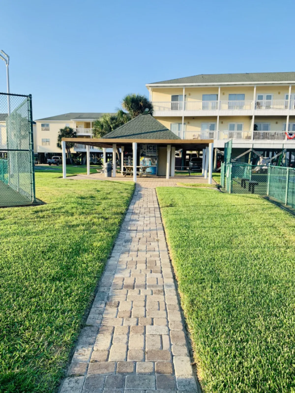 A brick pathway leads to a covered pavilion with picnic tables and vending machines. Behind the pavilion, tennis courts with green fencing invite guests for active play. The scene is framed by a three-story yellow condominium building and landscaped resort grounds.