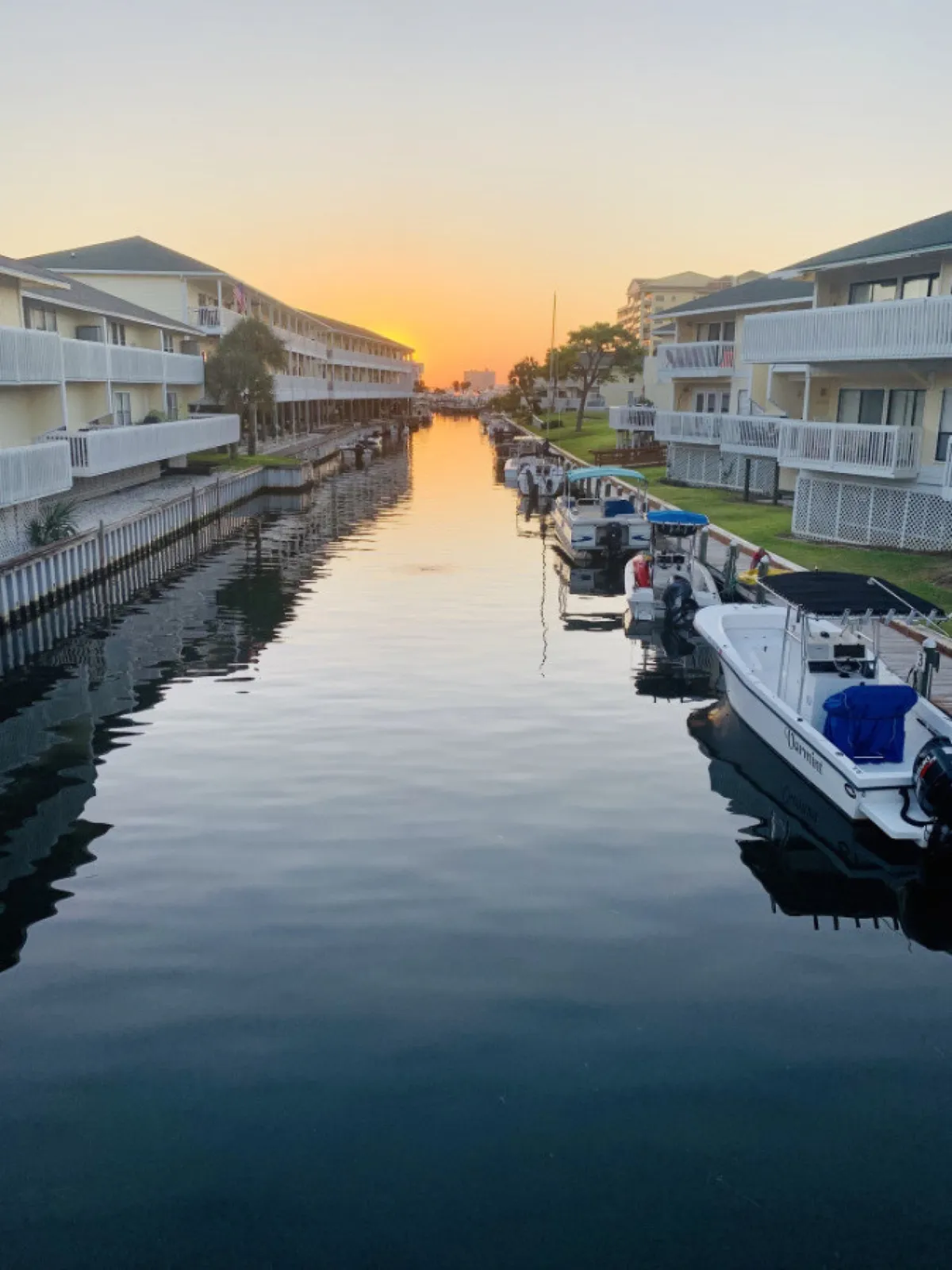 This image captures a tranquil canal lined with docked boats, flanked by modern condominium buildings with balconies. The warm sunset light reflects on both the water and the boats, evoking a sense of relaxation and the boating lifestyle. Balconies overlook the canal, emphasizing the connection between the resort and water-based activities.