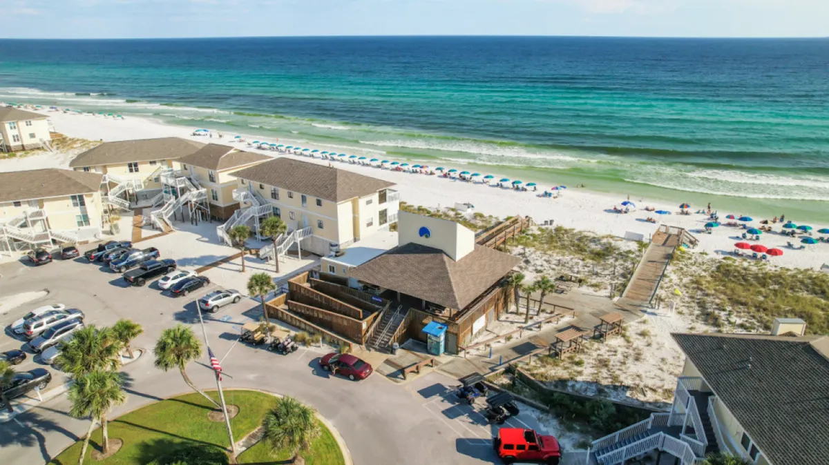 A broad view of the resort’s private white sandy beach, dotted with colorful umbrellas and lounge chairs, with the turquoise Gulf of Mexico stretching into the distance. The beachfront resort buildings with multi-level decks and a boardwalk leading directly to the beach are also visible, emphasizing the ease of beach access.